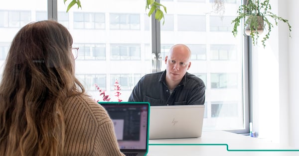 En man i svart skjorta sitter framför sin laptop och ser bekymrad ut, mitt emot honom sitter en kvinna i en brun stickad tröja framför sin laptop. Bakom mannen syns en företagsbyggnad.