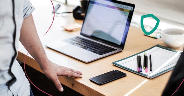 En bild på en person som står vid sitt skrivbord. På bordet står det en dator, en telefon och ett blogg med pennor. Personen har ett antivirusprogram.